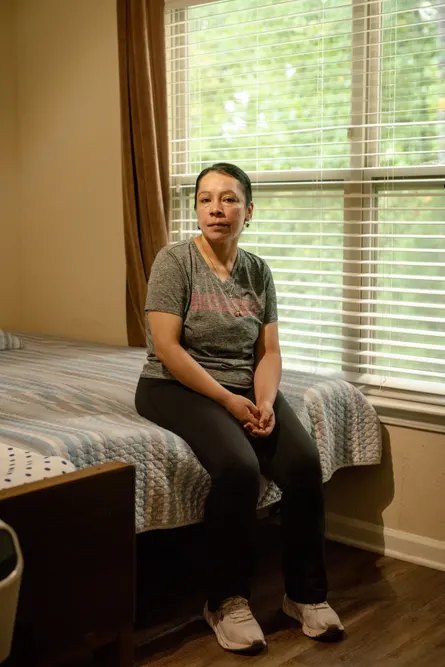 woman sits on edge of bed next to a window facing trees, her hands clasped