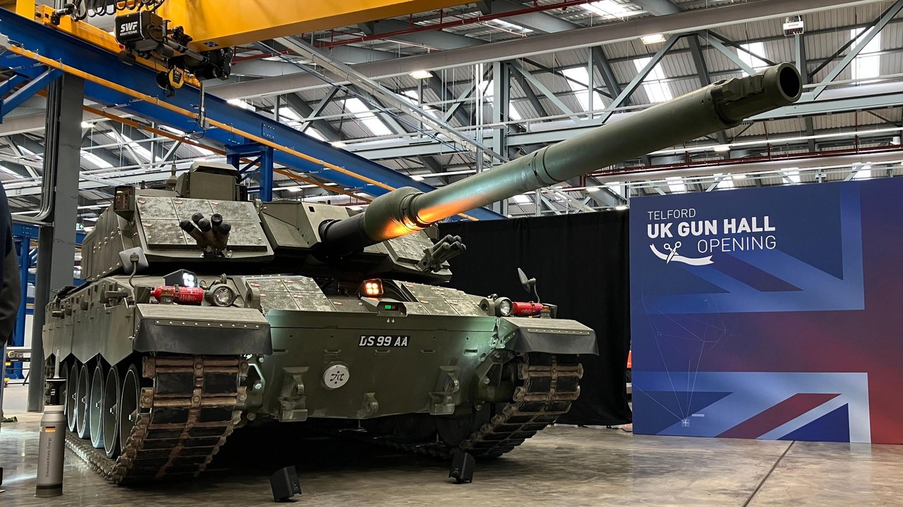 A green tank is in the middle of a large warehouse building as part of a display. There is a display board next to it with a union flag background and it says "Telford UK Gun Hall Opening".