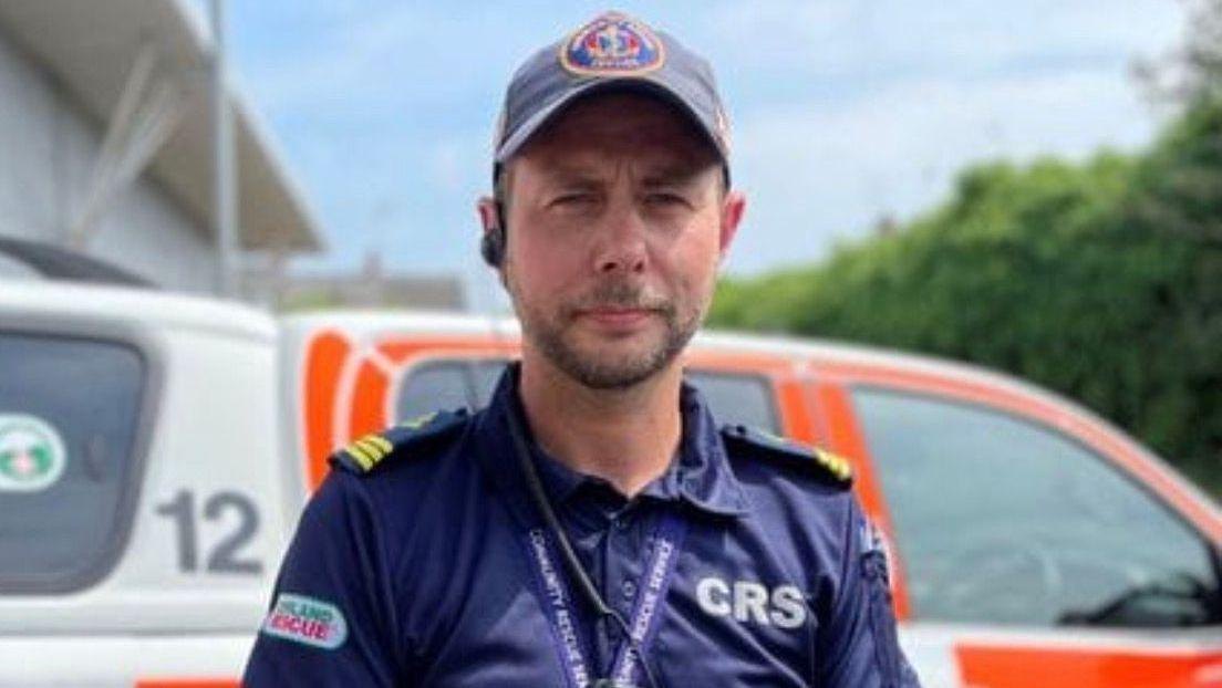 A man in a navy cap wearing a navy shirt. He has a brown beard. The photo's qualitiy is not good.