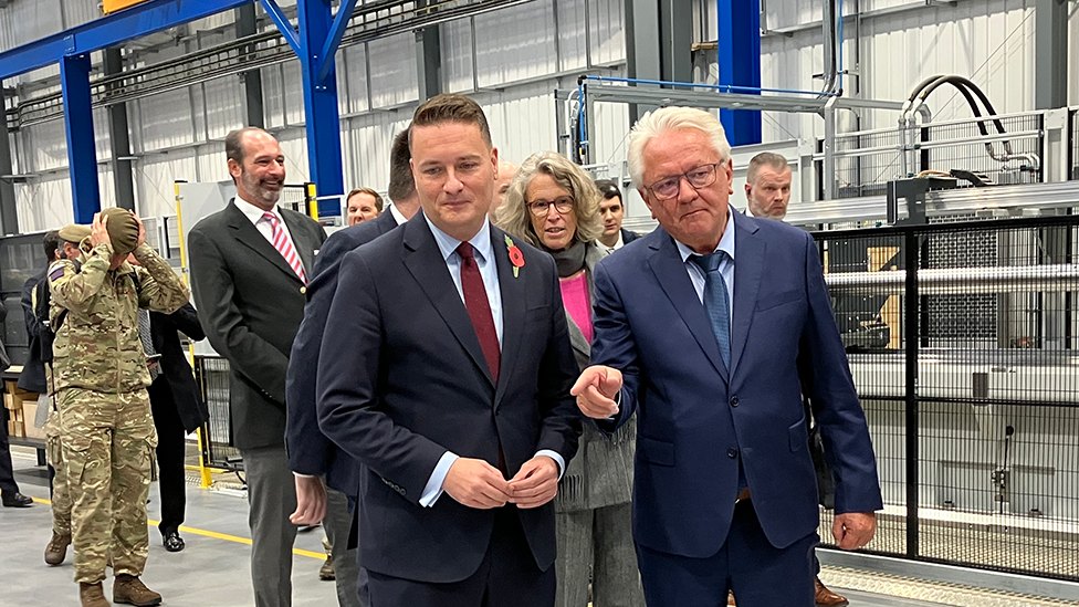 Wes Streeting and another man talk together on a factory floor. There are others in the background, including members of the armed services in uniform