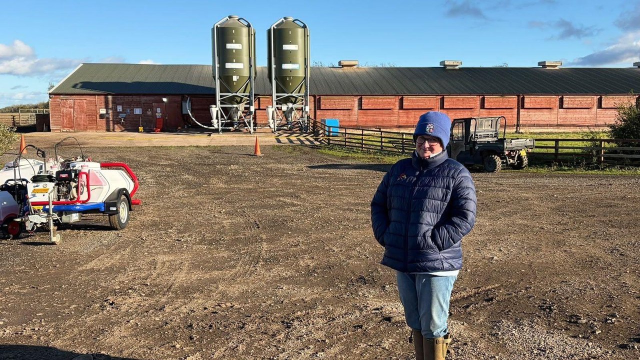 Catherine, dressed in a blue puffer jacket, jeans, woolly hat and green wellies, stands in her farm yard. Behind her stands the red brick barn where her flock were kept.