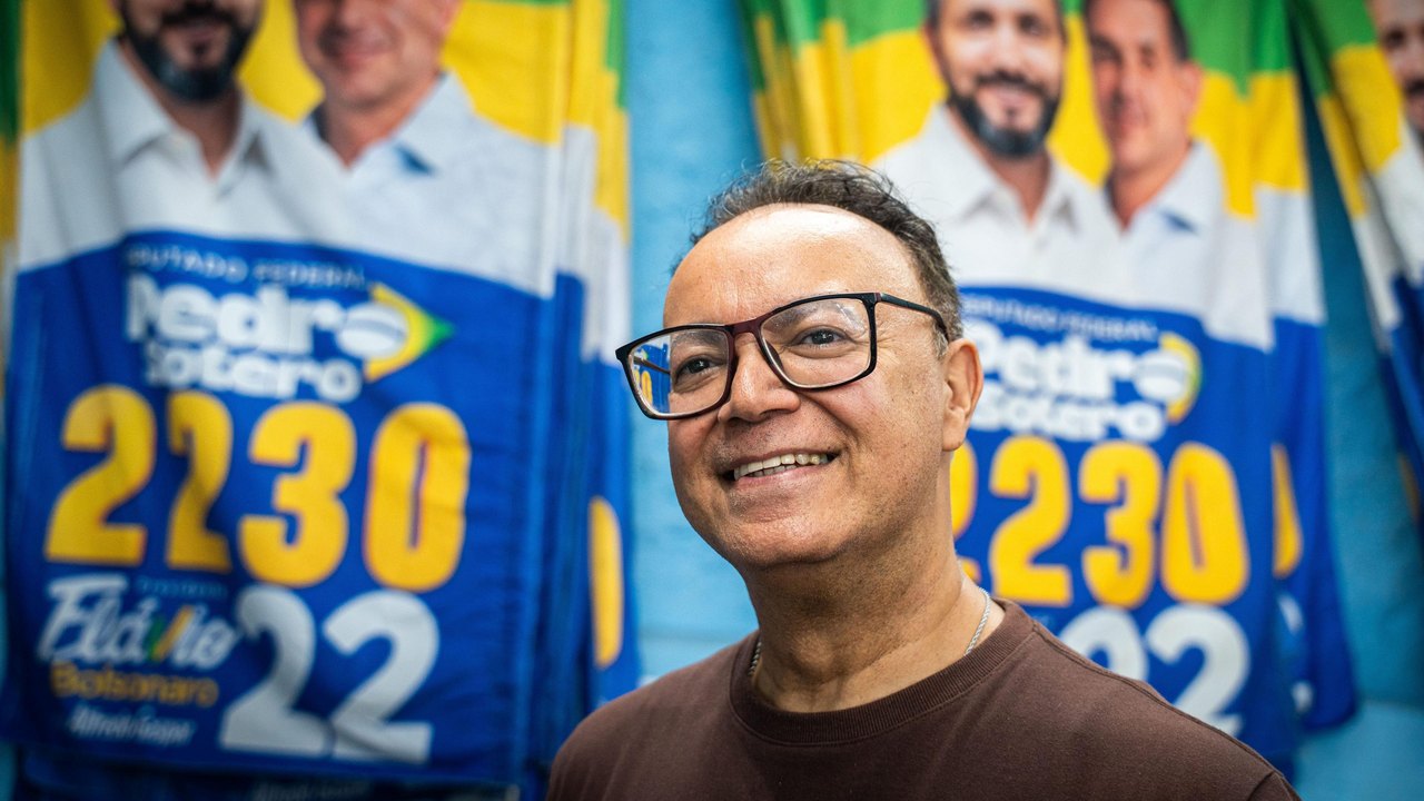 Man in a brown shirt and glasses standing front of Bolsonaro banners.