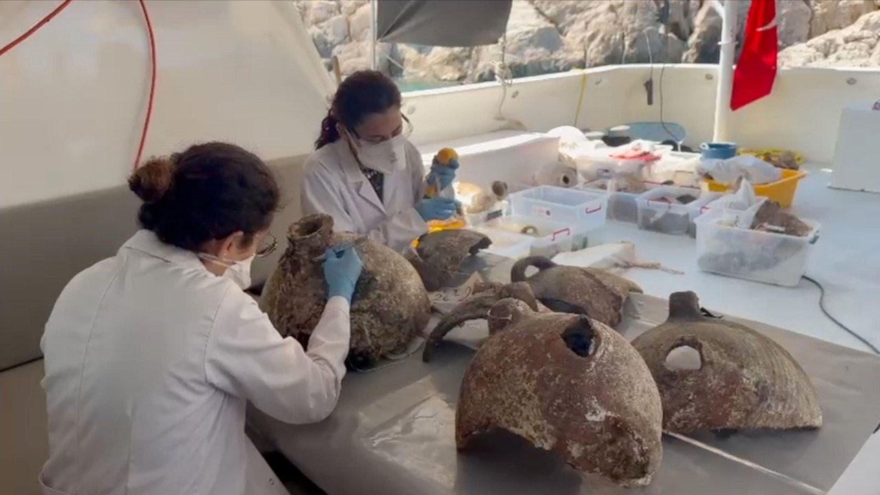 Two people in lab coats and wearing blue gloves and masks are examining some of the amphorae that have been brought to the surface. They are sitting at a table at the back of a dive boat and are in the shade under the boat's roof. The person on the left has her hair tied up and is looking at an intact amphora. She is gently and carefully scraping the surface close to the neck with a scalpel. There are 4 other partial amphorae on the table. Another person who sits slightly behind and to the left,