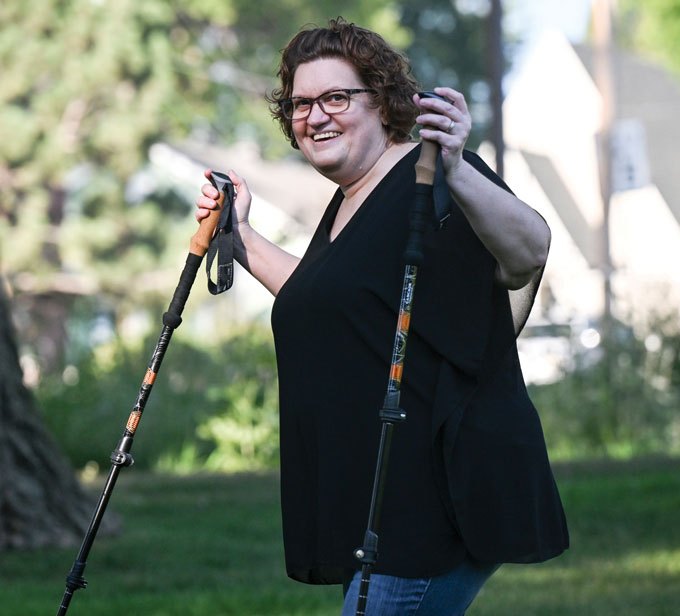 A smiling person with short, curly brown hair and glasses stands outdoors, holding a trekking pole in each hand. They wear a black top and blue jeans, with trees and buildings blurred in the background.
