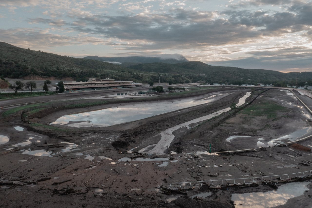 An elevated view shows a mud-caked, flooded horse-racing track surrounded by standing water and mountain ridges under a cloudy sky.