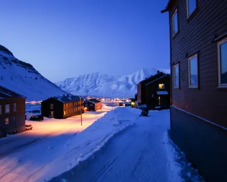 Snow-covered wooden houses line a road at dusk in Longyearbyen, Svalbard, mountains behind