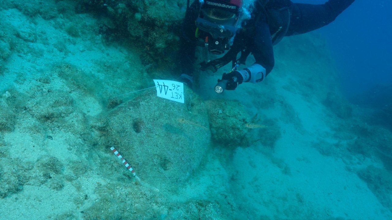 The picture is taken underwater and has a deep blue green colour. In the centre is a large shield-shaped rock with two holes a little way apart at one end and another one at the other. It is about 1m across. This is one of three ship's anchors found at the site. A piece of paper is stuck to the anchor with "KET 26 - 44" written on it. An archaeologist's red and white measuring stick is lying on the seabed at the end with the 2 holes. To the right of the stone anchor, and slightly above, where th
