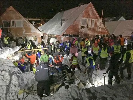 People digging into a pile of snow at night by several damaged houses