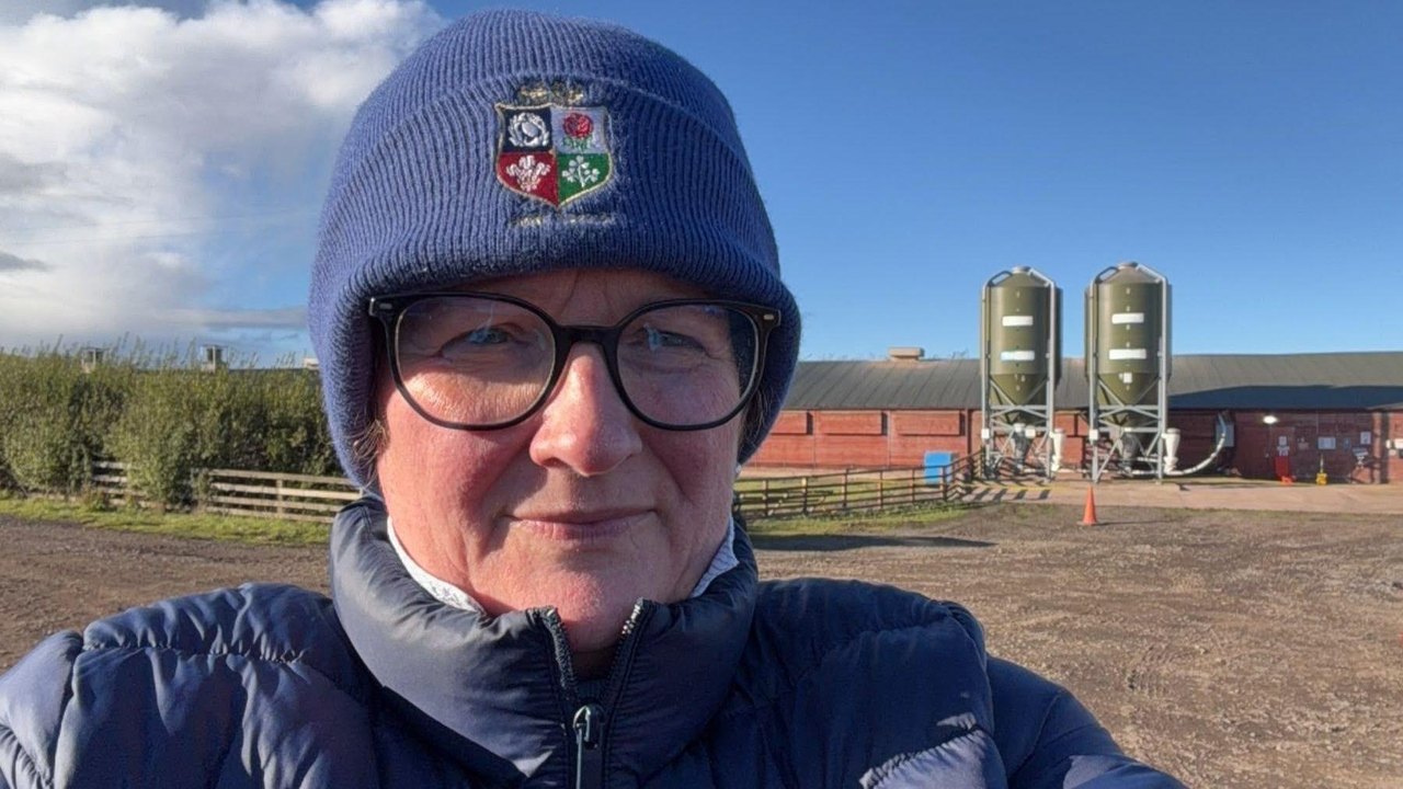 In a selfie-style head-and-shoulders shot, Catherine is wearing glasses, a blue woolly hat and matching puffer jacket. In the background is the farmyard and a long poultry barn.