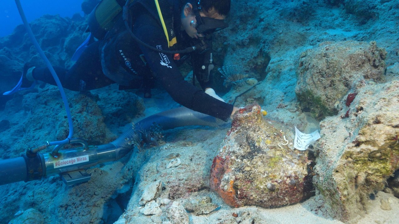 A diver on the top left hand side of the picture is working underwater to uncover an amphora. They are wearing a black wet suit with some white writing on it and a yellow stripe. They have black goggles. A long tube, like the sort you find on a vacuum cleaner, is being used to remove the sand that has been covering the ship's cargo. In the foreground and slightly to the right is an amphora. It is mostly covered in a sort of multicoloured rocky crust and it is sitting on a small ledge but some