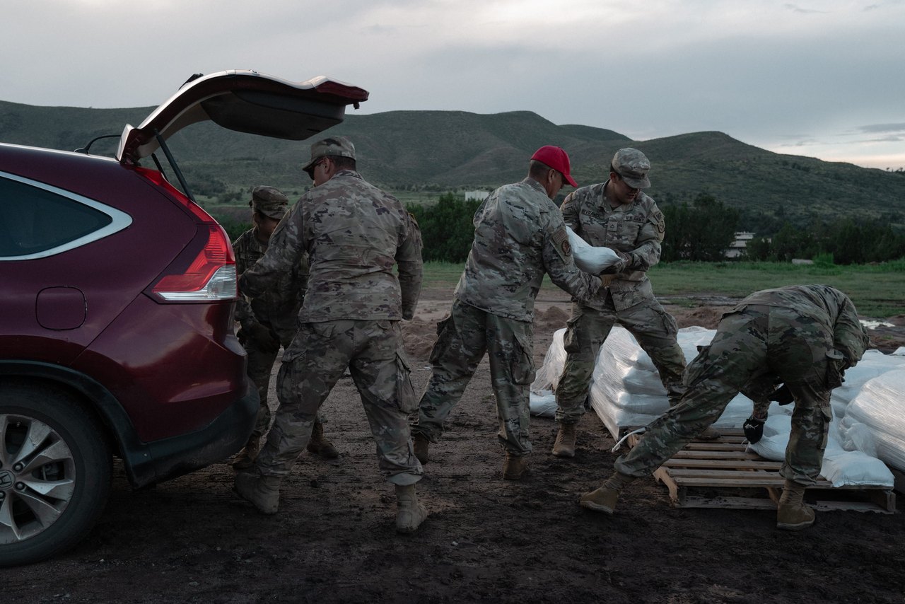 National Guard service members load white sandbags from wooden pallets into the open trunk of an SUV ahead of flood preparations.