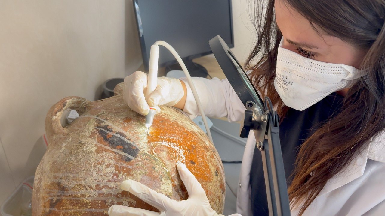 A close up view of an amphora in the laboratory on the ship. The handle on the side of the pot is poking out to the left. A woman with long brown hair, wearing a white lab coat and a face mask, is working on the surface of the amphora with a small electric tool. It's white and is about 10cm long with a light on the end, she is holding it in her right hand and supporting the amphora with her left. The area that she has carefully cleaned is a brown/orange colour and there are some black patterns t