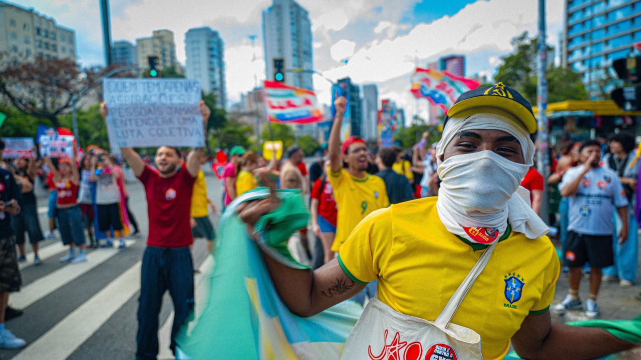 A young man covering his face in a white cloth wearing a yellow Brazil football shirt waving a Brazil flag. In the Background protesters hold up flags and signs.