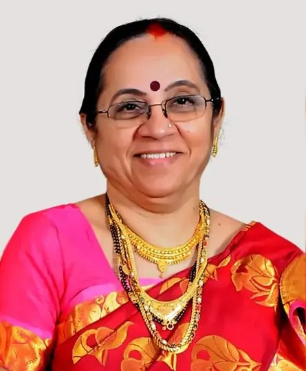 A smiling older Indian woman, wearing glasses and a red sari, with layered gold necklaces
