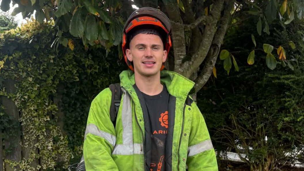 Callum Kirk is wearing a orange helmet, a yellow his-vis jacket, with a black t-shirt underneath. He has equipment in his hand. There is a tree and a bush in the background.