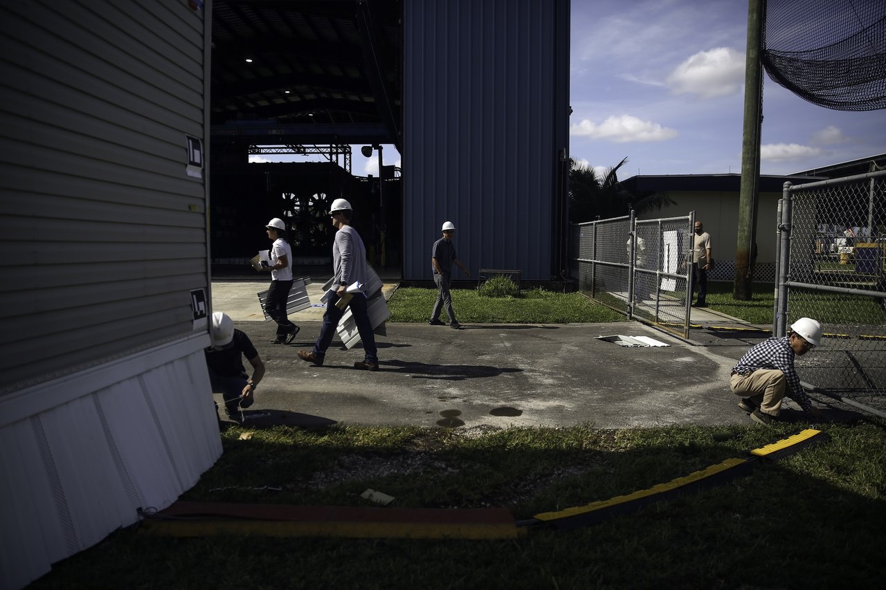 a group of people wearing hard hats walk around a corner near a wind testing site for manufactured housing