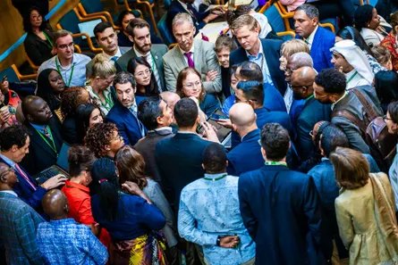 A cluster of about 50 people from all around the world standing close together in a hall as they discuss something