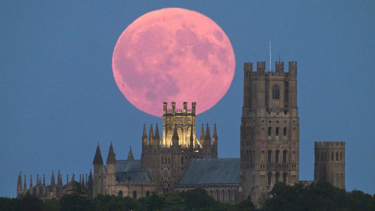 A large reddish moon hangs in the dark blue sky behind Ely cathedral