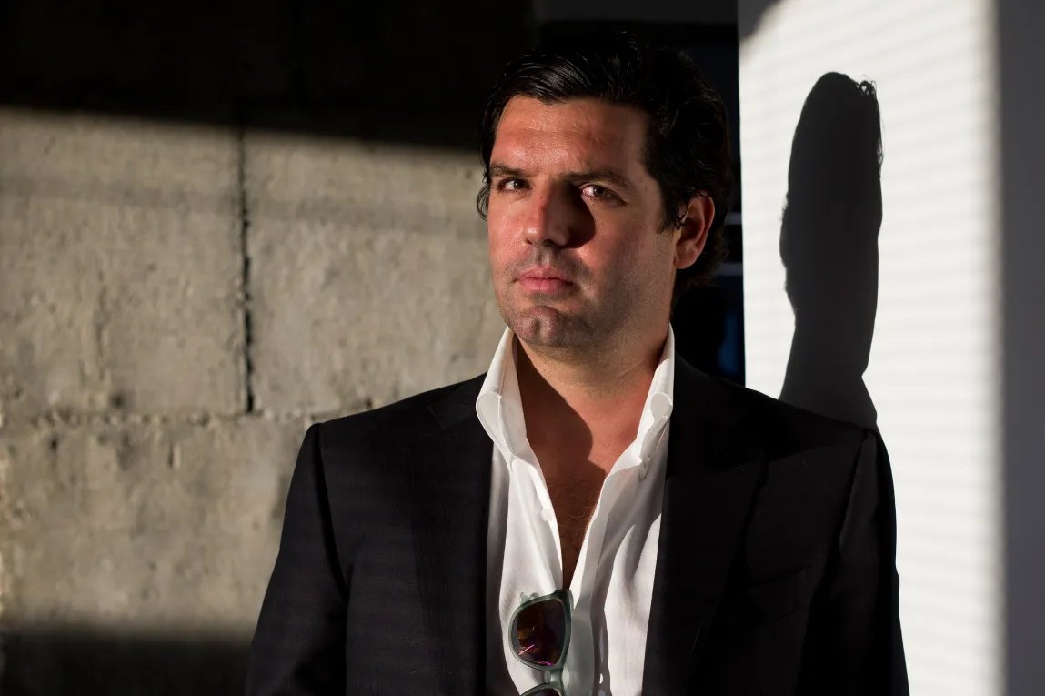 A man in a sportscoat looks at the camera. He is lit dramatically and casts a shadow on the wall behind him.