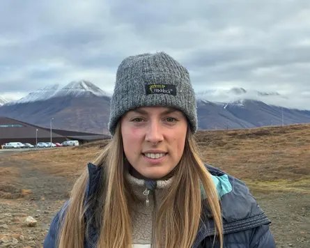 A young white woman stands outdoors in a beanie and jacket, with snow-capped mountains behind her