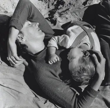 A mother lies on sand with eyes closed, cradling her baby against her chest