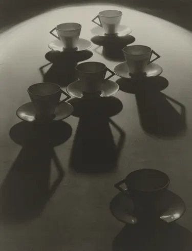 Six teacups and saucers cast long dramatic shadows on a curved surface, in black and white
