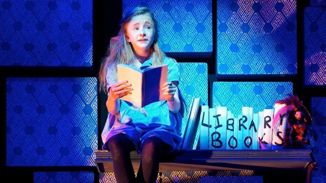 The 2011 child actor playing Matilda sits on a wooden shelf with library book props next to her on stage. She is holding a book and has a blue tinged set behind her.