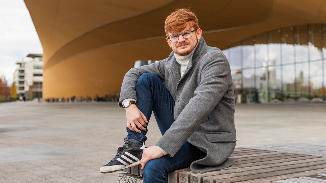 Owein Iveson sits outside, wearing a grey coat, blue jeans and trainers