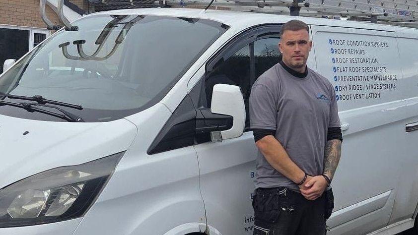 Charles Howard is standing with a grey top on, with black trousers and black shoes. He is standing infront of a white van.